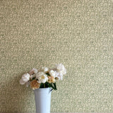 A vase of flowers stands in front of a wall papered in a painterly leaf print in olive and black on a cream field.