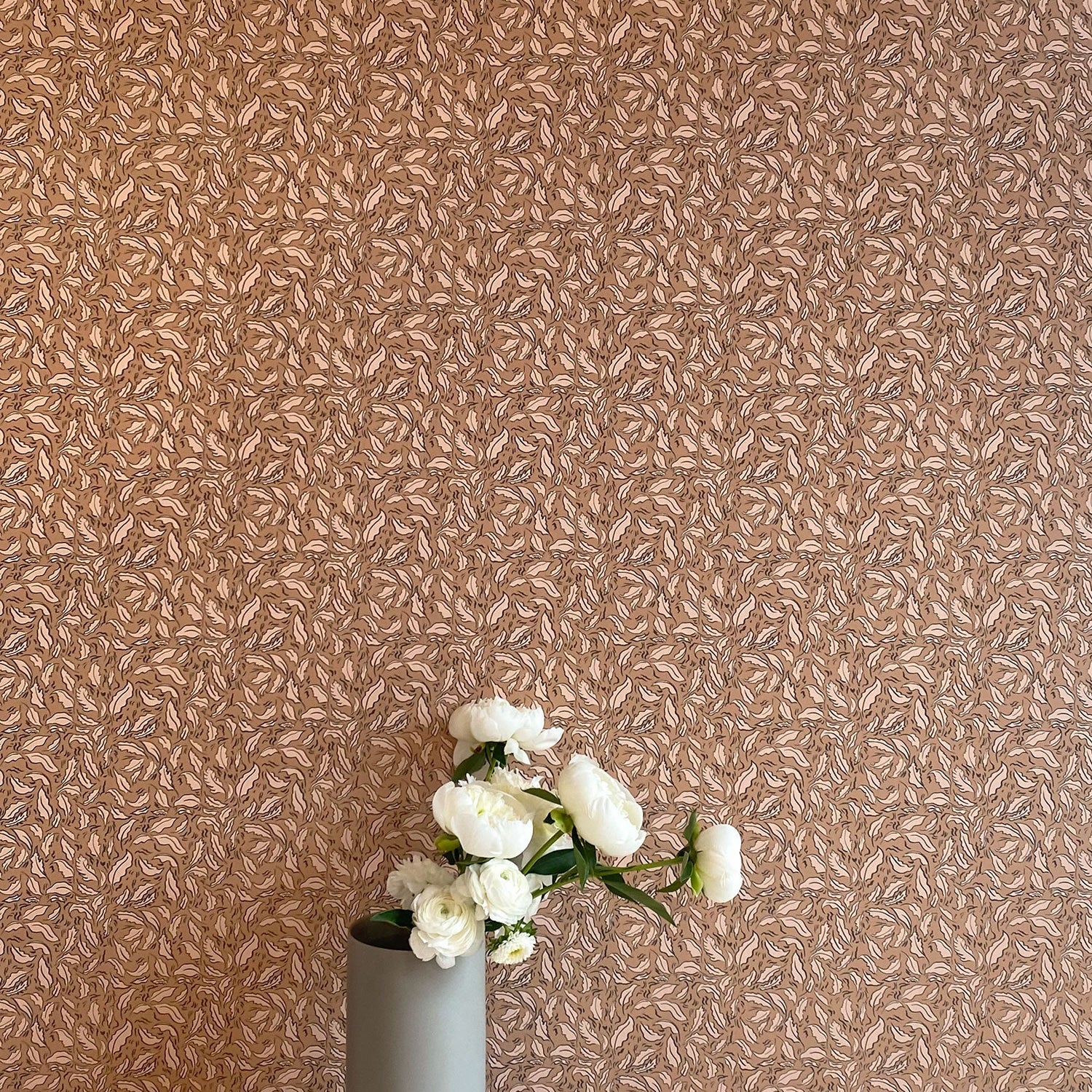 A vase of flowers stands in front of a wall papered in a painterly leaf print in cream and black on a brown field.