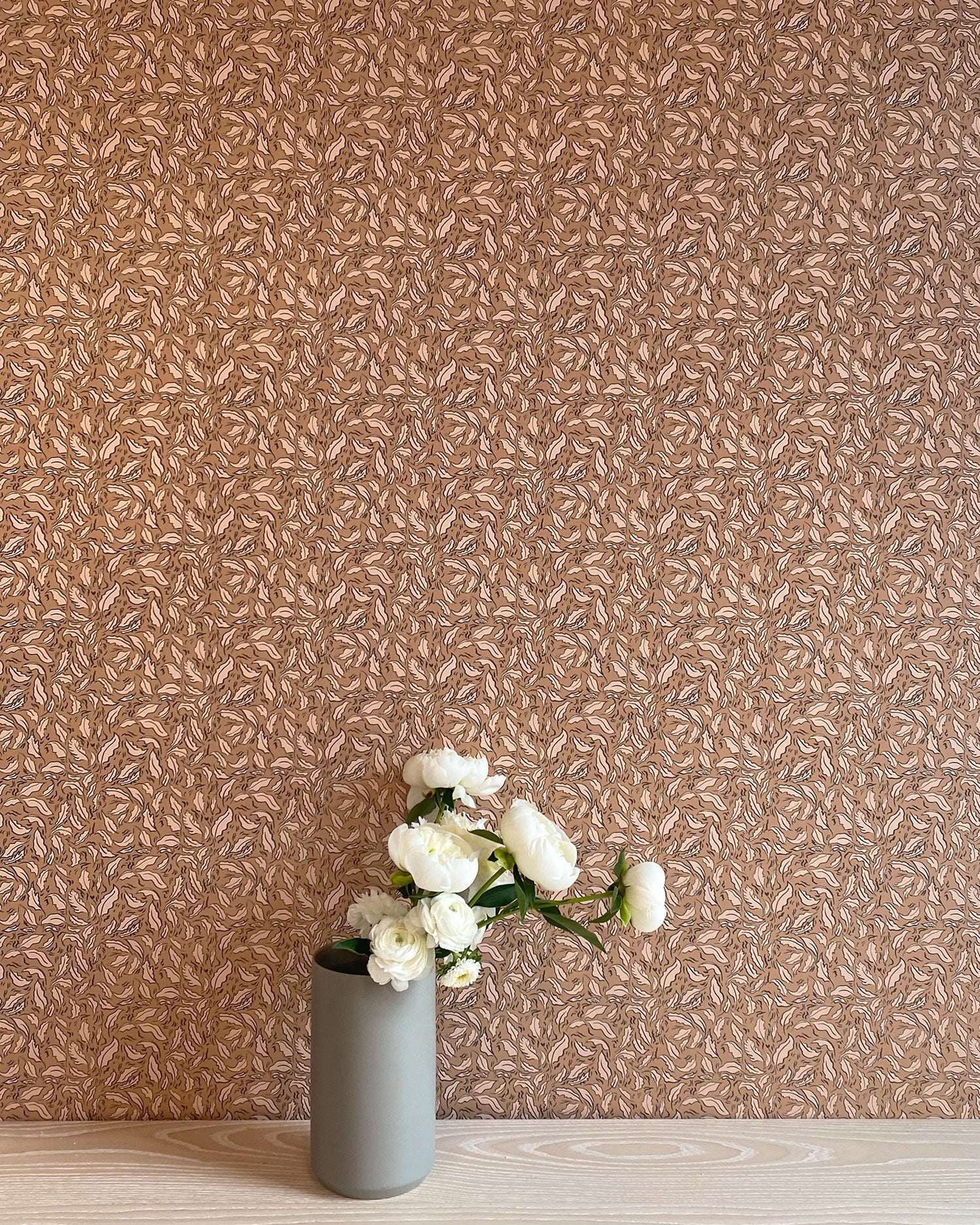 A vase of flowers stands in front of a wall papered in a painterly leaf print in cream and black on a brown field.