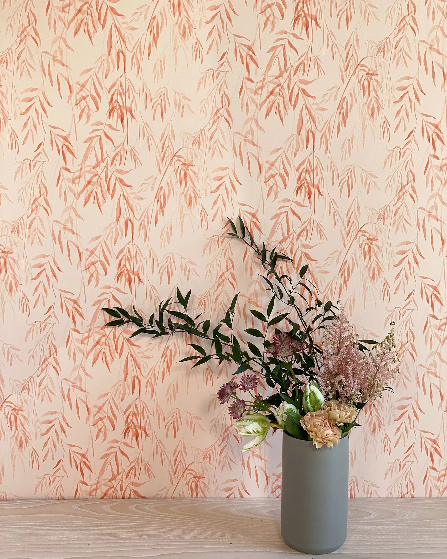A vase of flowers stands in front of a wall papered in a painterly willow leaf print in coral on a cream field.