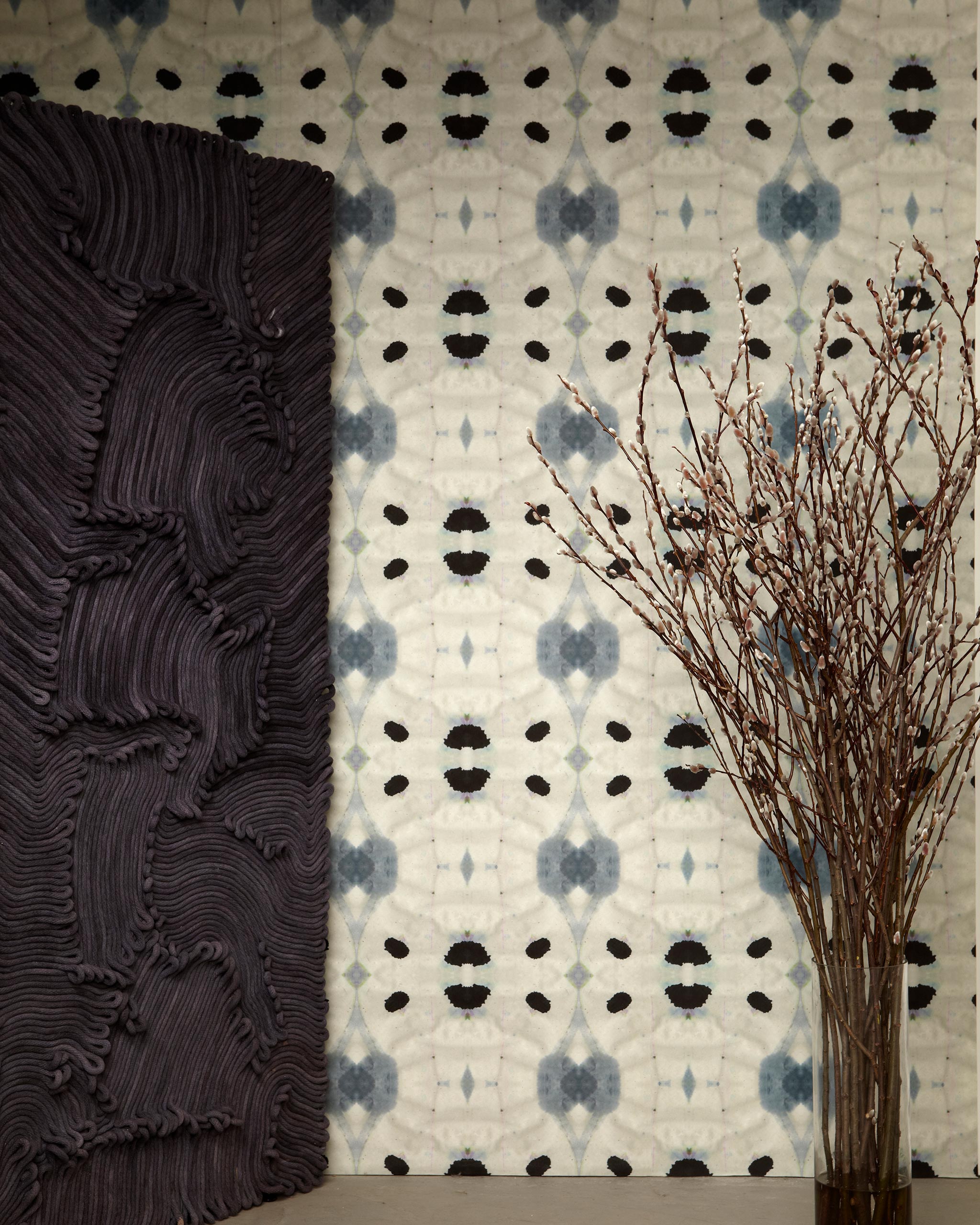Flowers and a black room divider stand in front of a wall papered in a watercolor ink blot print in charcoal, black and tan.