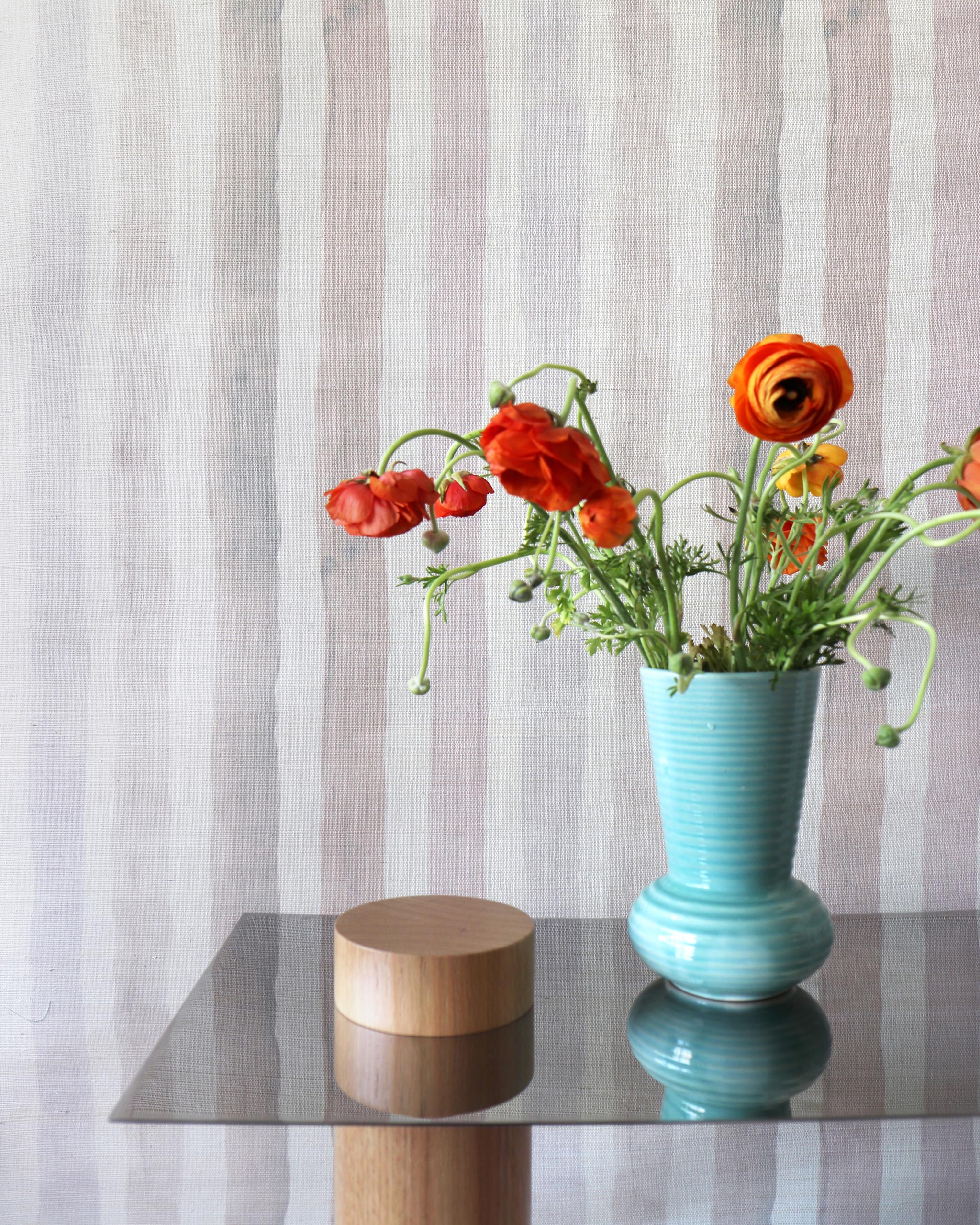 An end table with flowers stands in front of a wall papered in a painterly stripe print in pink, gray, cream and white.