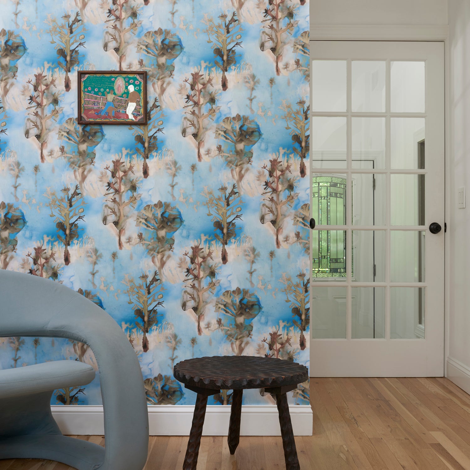Living room interior with blue accent chair against wallpaper in watercolor tree print in shades of tan and gray on a blue field.