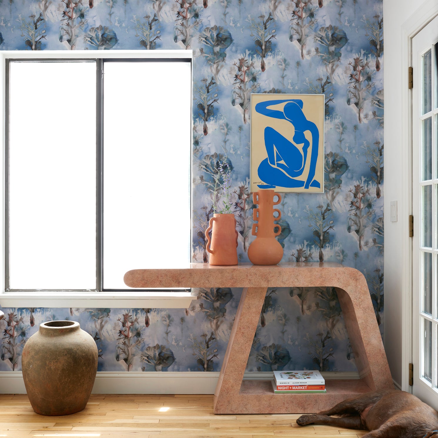 Living room interior with sculptural table and props against a watercolor tree print in shades of turquoise and grey on a blue field. 
