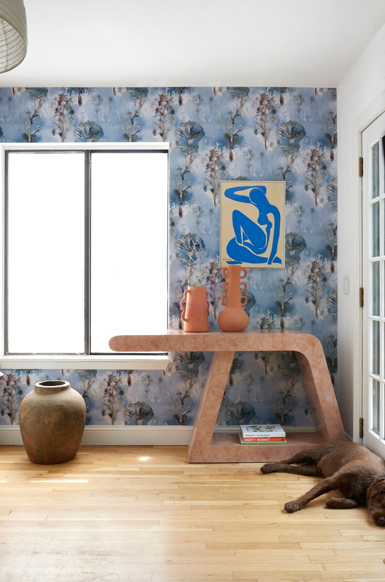 Living room interior with sculptural table and props against a watercolor tree print in shades of turquoise and grey on a blue field. 