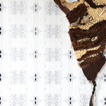 Wallpaper in a minimalist ikat pattern in shades of gray on a white field with a handwoven textile sculpture and woven bench. 