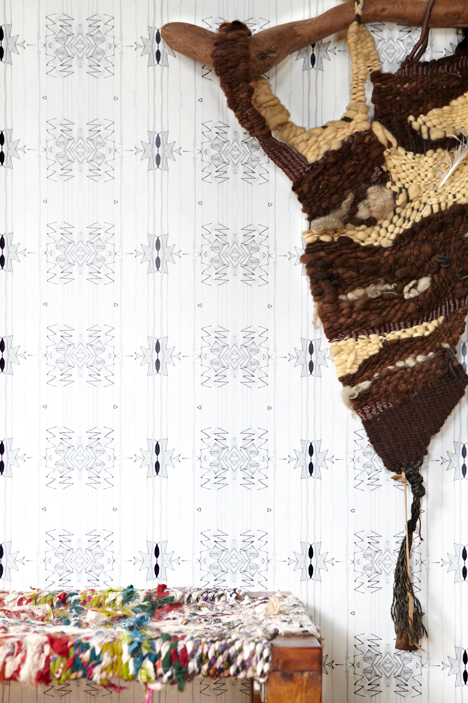 Wallpaper in a minimalist ikat pattern in shades of gray on a white field with a handwoven textile sculpture and woven bench. 