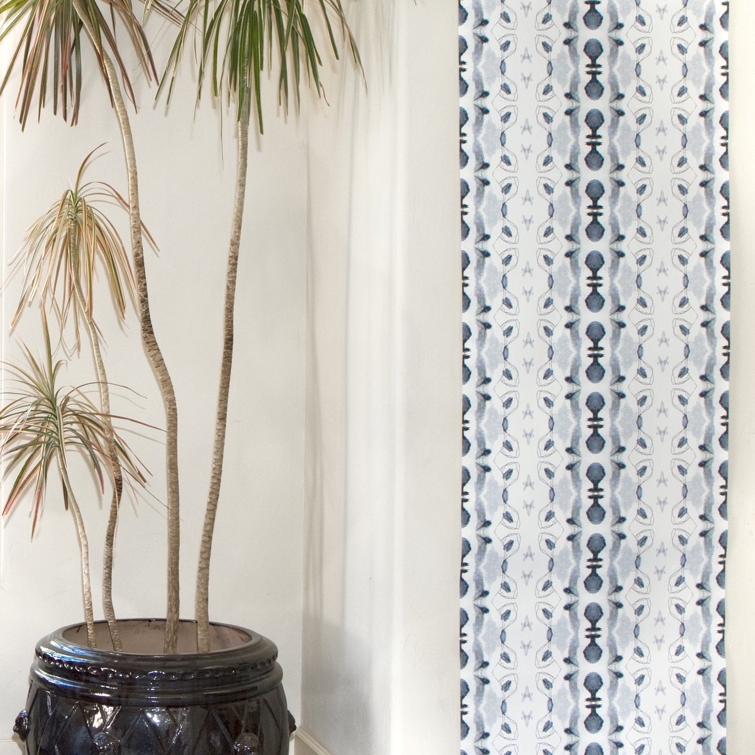 Wallpaper roll in a watercolor ikat stripe print in shades of blue and black on a white field draped against a wall with a potted plant.
