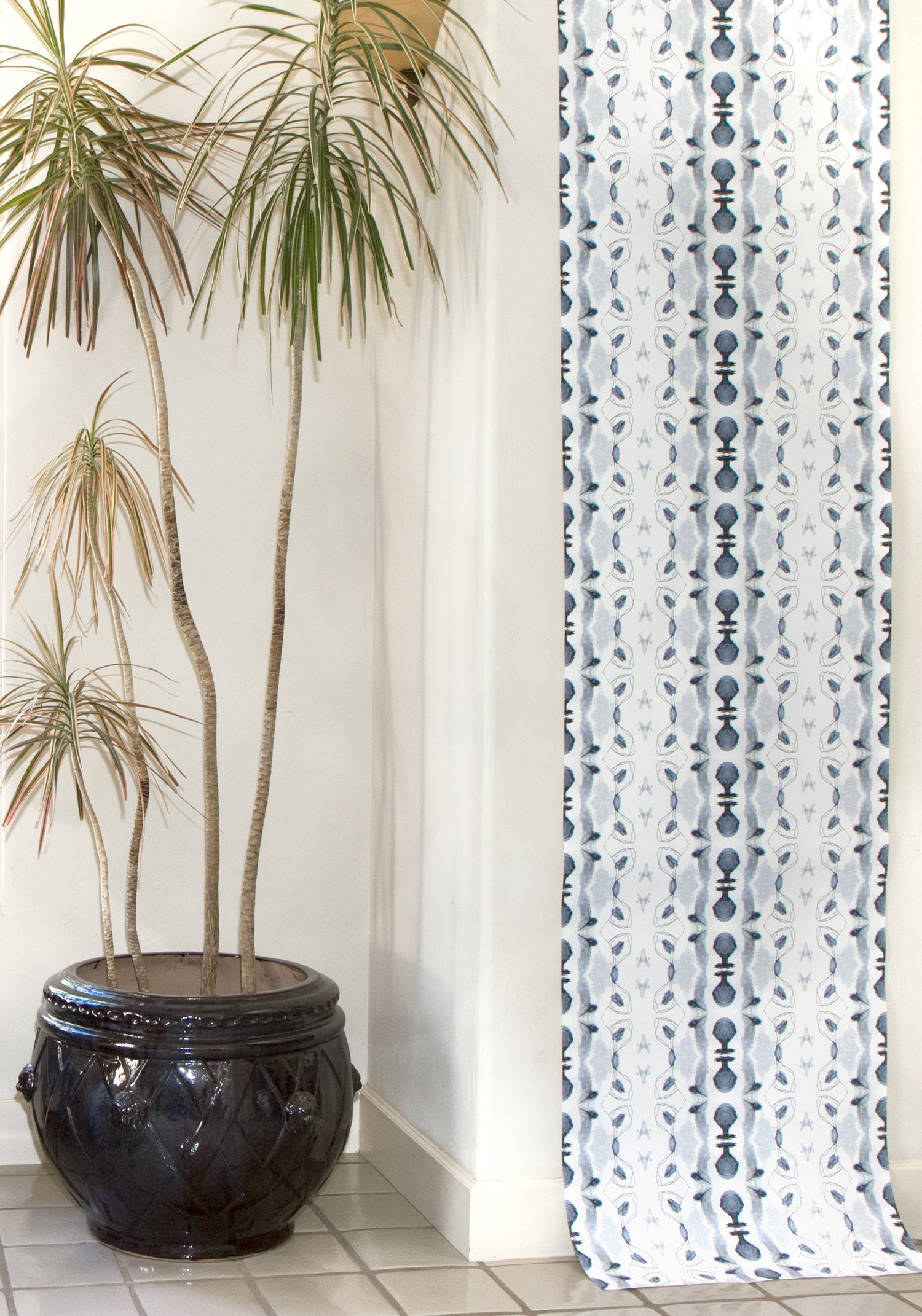 Wallpaper roll in a watercolor ikat stripe print in shades of blue and black on a white field draped against a wall with a potted plant.