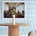 Wallpaper install in a painterly stripe print in blue on a white field, witha framed photo, wooden dining table and flower vase. 