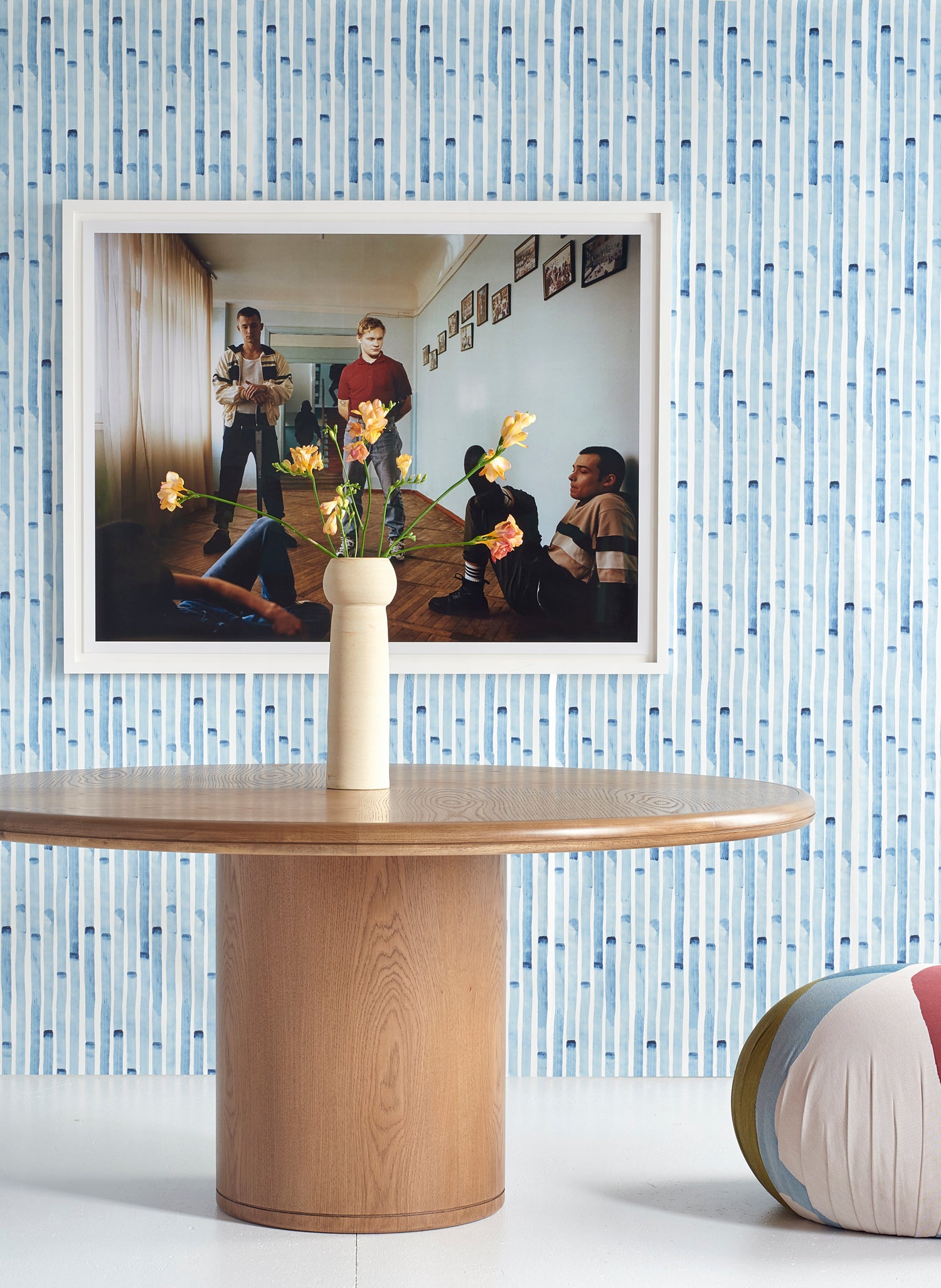 Wallpaper install in a painterly stripe print in blue on a white field, witha framed photo, wooden dining table and flower vase. 