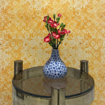 Small round table with a blue polka dot vase and pink flowers against a yellow floral wallpaper.
