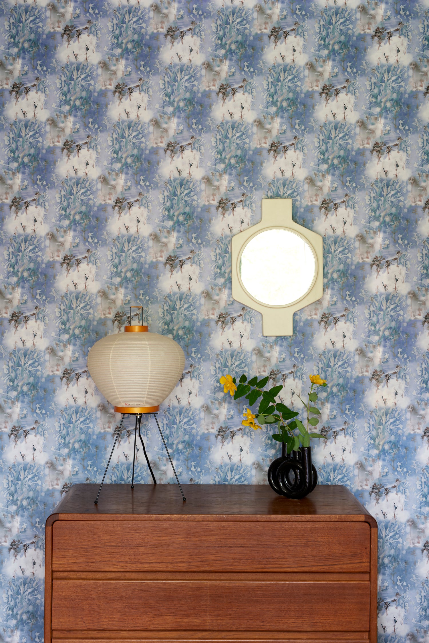Wooden credenza with paper lantern and props against in a painterly tree print in blue and grey. 