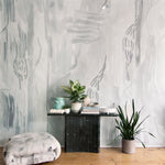 Living rooms scene with pedestal table, ottoman and plants against a watercolor wallpaper mural in shades of grey. 