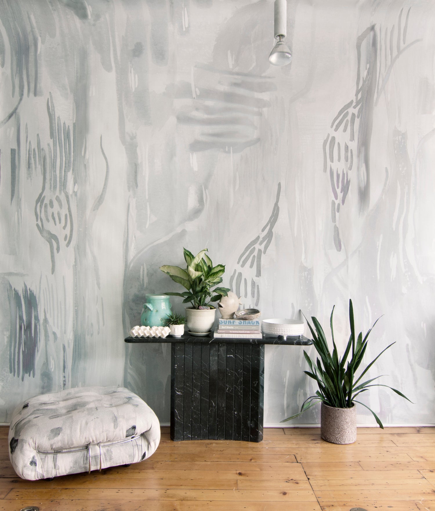 Living rooms scene with pedestal table, ottoman and plants against a watercolor wallpaper mural in shades of grey. 