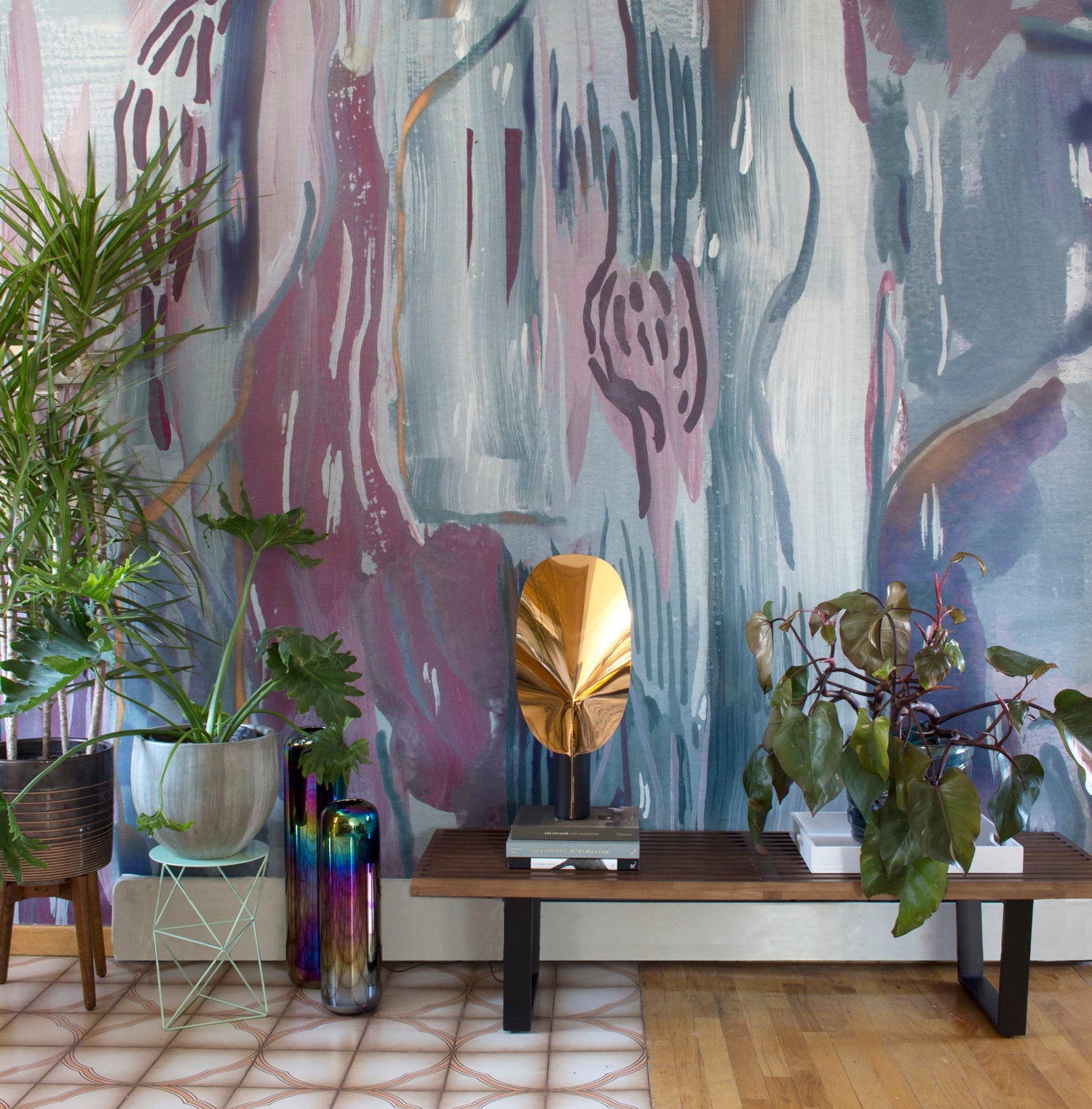 Living rooms scene with wooden bench and plants against a watercolor wallpaper mural in purple and teal.  