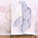 Wallpaper rolls in a in a watercolor watercolor print in shades of gray on a white field against a wall in the same pattern in pastel pink. 