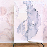 Wallpaper rolls in a in a watercolor watercolor print in shades of gray on a white field against a wall in the same pattern in pastel pink. 