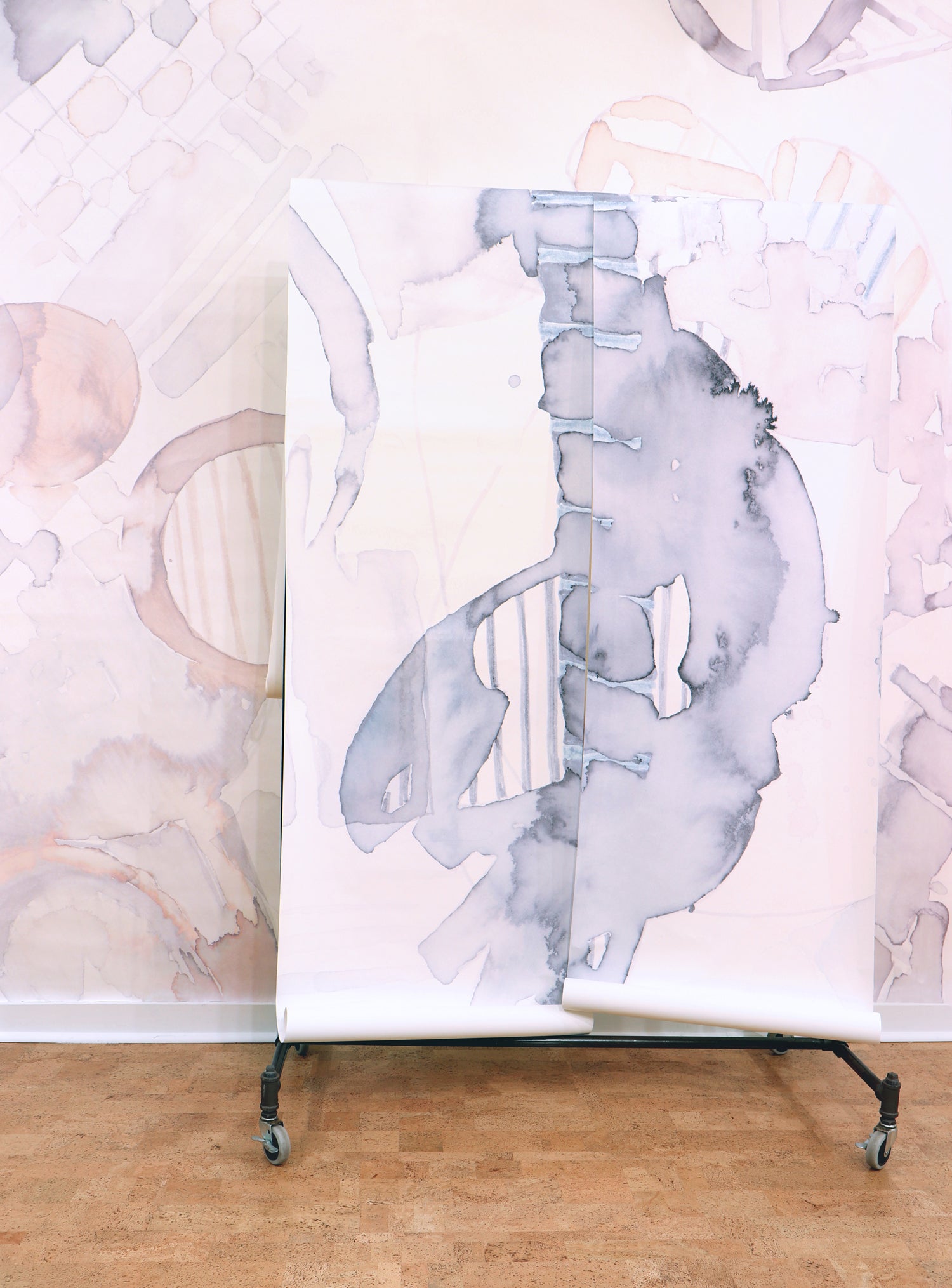 Wallpaper rolls in a in a watercolor watercolor print in shades of gray on a white field against a wall in the same pattern in pastel pink. 