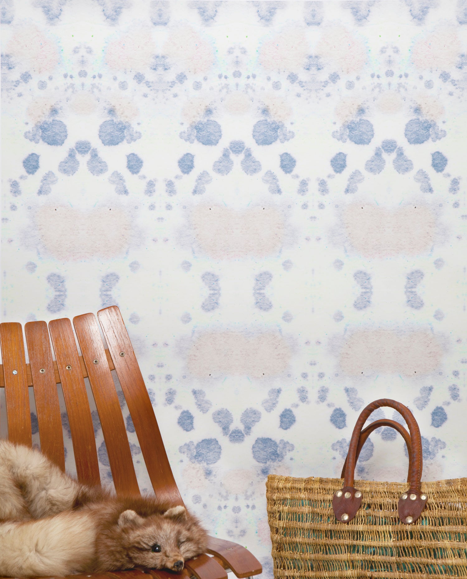Wallpaper install detail in a watercolor inkblot print in pastel pink and purple on a cream field with a wooden slat chair and woven bag. 