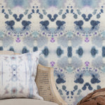Wallpaper install vignette with a wicker side chair against watercolor inkblot print in shades of blue and purple on a cream field.