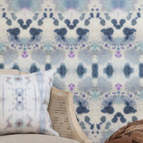 Wallpaper install vignette with a wicker side chair against watercolor inkblot print in shades of blue and purple on a cream field.
