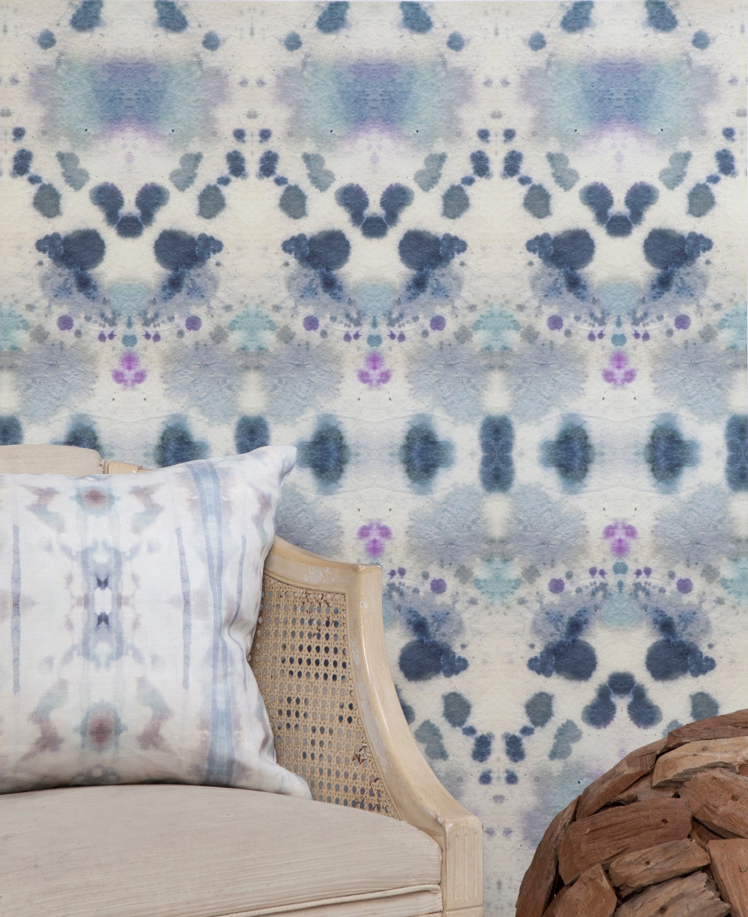 Wallpaper install vignette with a wicker side chair against watercolor inkblot print in shades of blue and purple on a cream field.