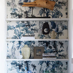 Built in white book case styled with books and props against a blue watercolor palm wallpaper at the back wall.