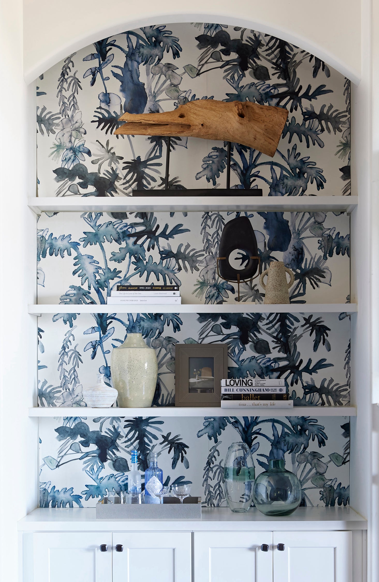 Built in white book case styled with books and props against a blue watercolor palm wallpaper at the back wall.
