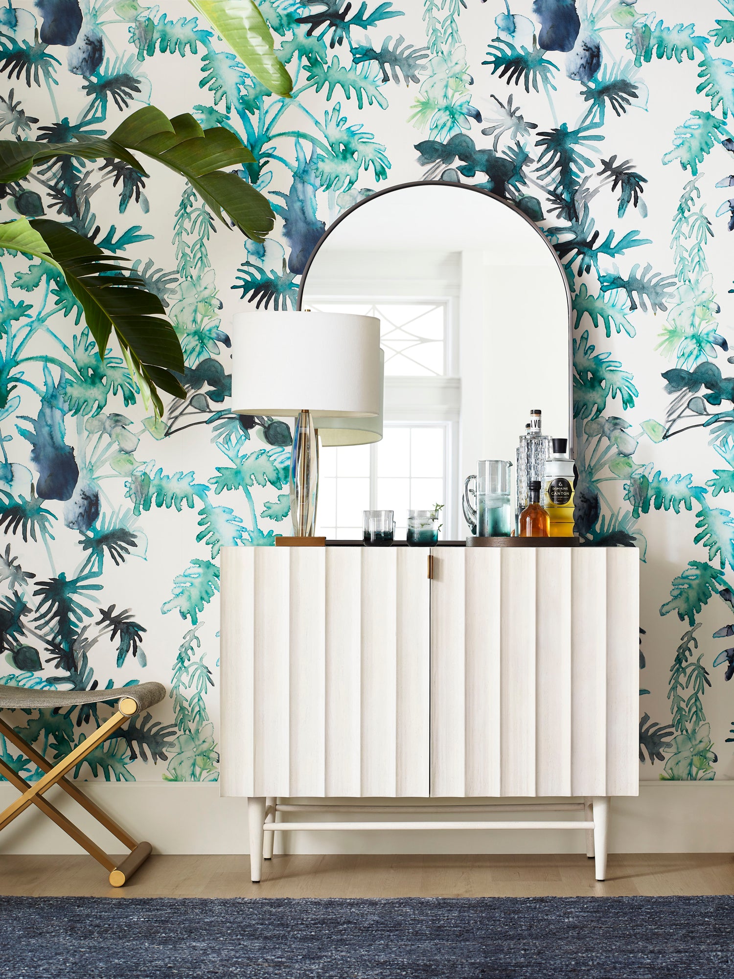 White fluted credenza with bottles and mirrors against a green palm frond wallpaper. 