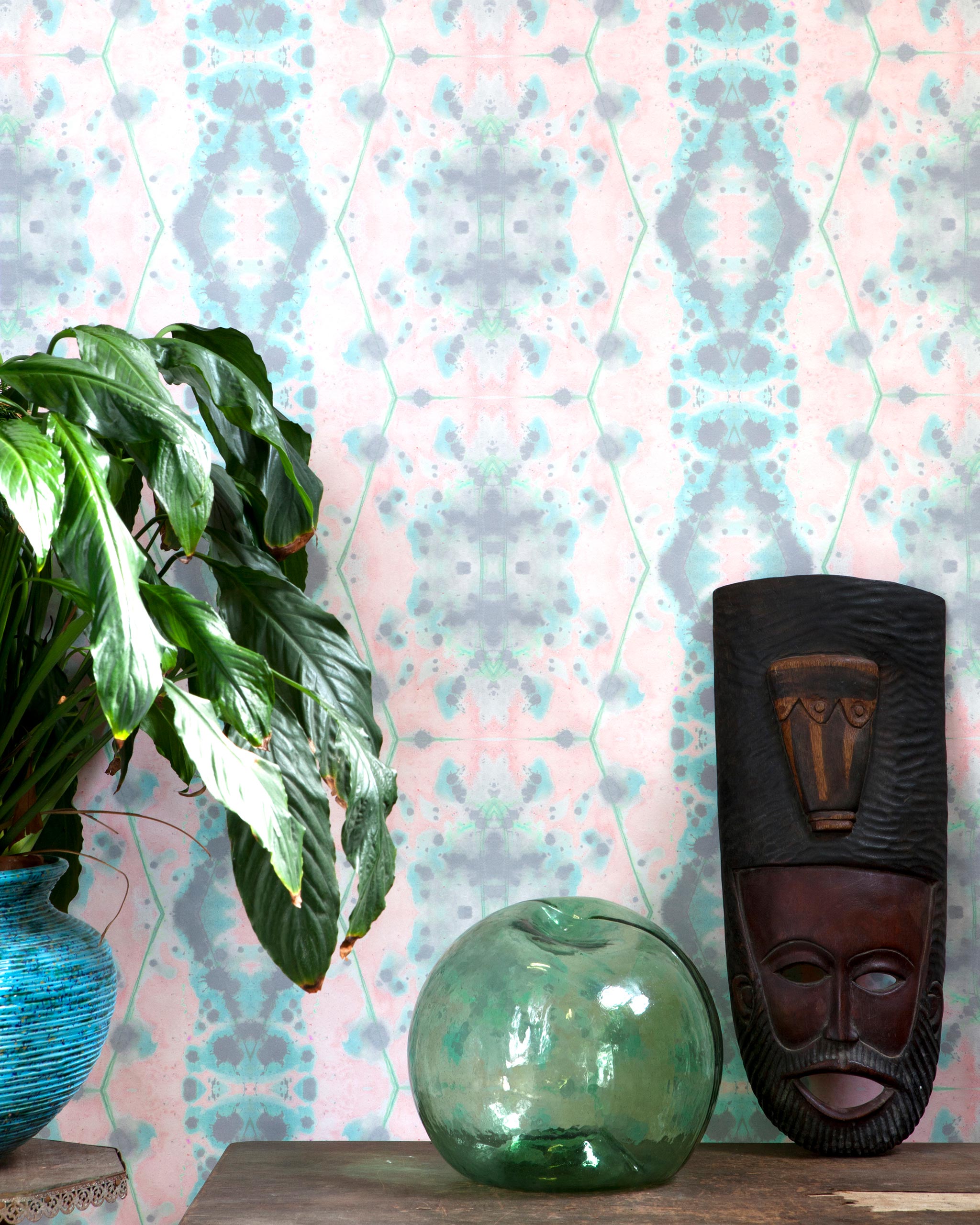 A plant and some knick knacks stand in front of a wall papered in a painterly gometric stripe in gray, blue, green and pink.