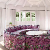 A large round sofa upholstered in a painterly tree print in purple, green and brown stands in the middle of a sunny room.