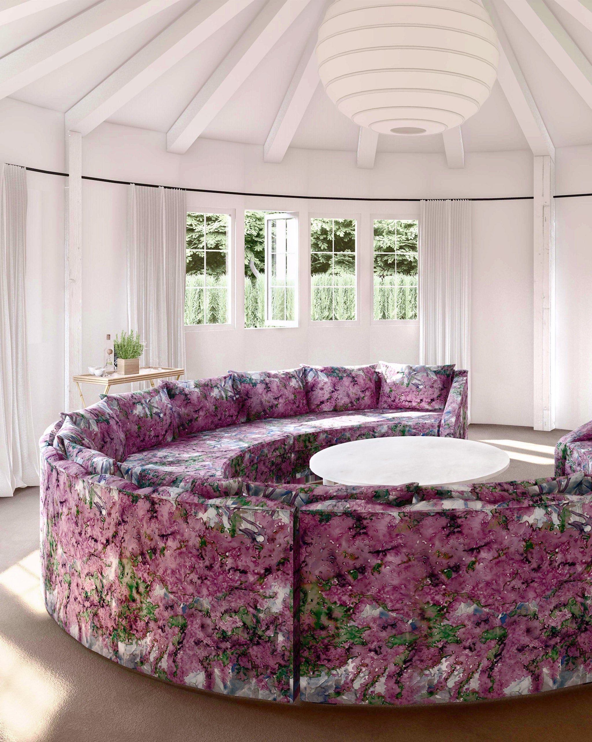 A large round sofa upholstered in a painterly tree print in purple, green and brown stands in the middle of a sunny room.