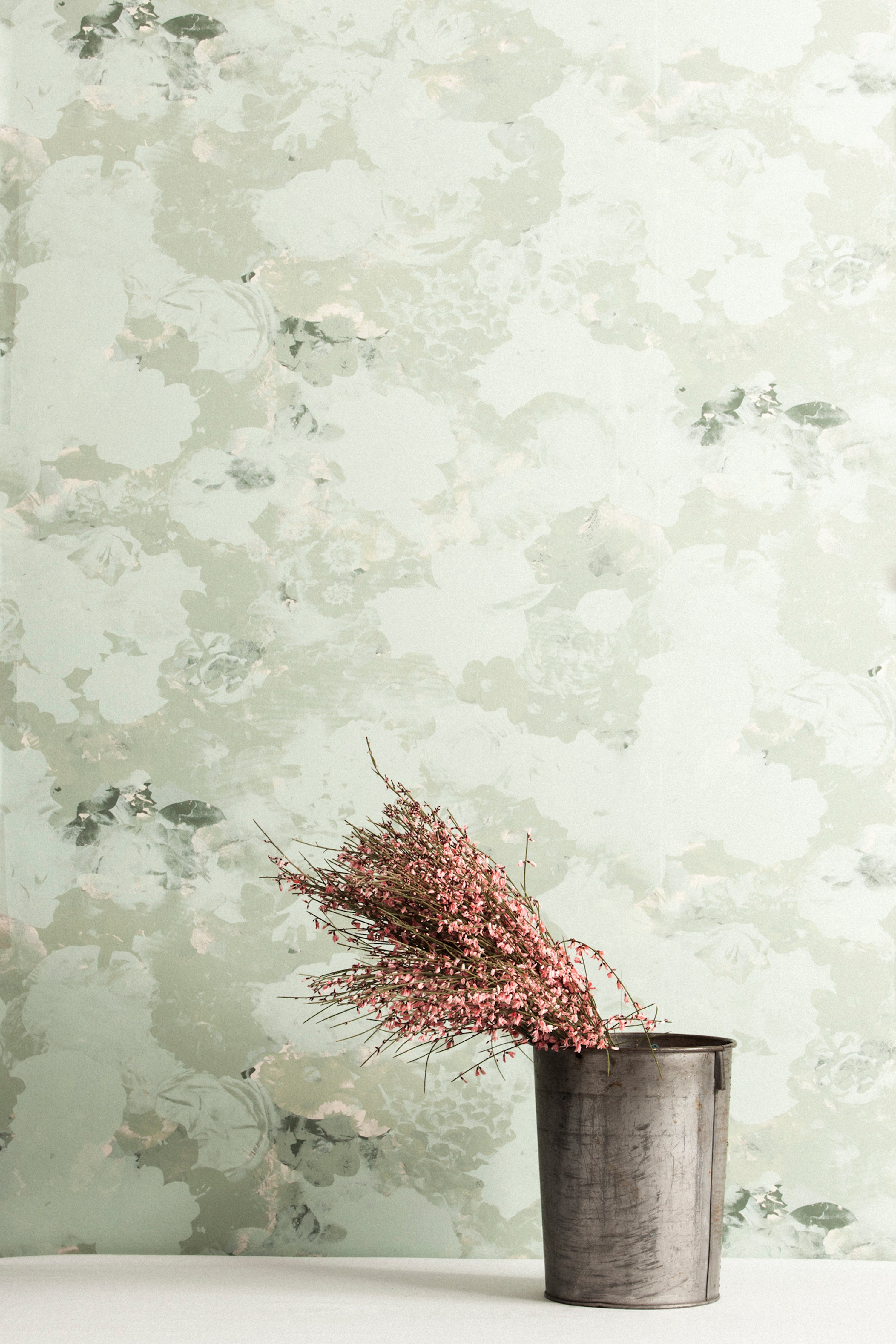 A vase of flowers stands in front of a wall covered in a dense photorealistic floral print in gray and pastel green.