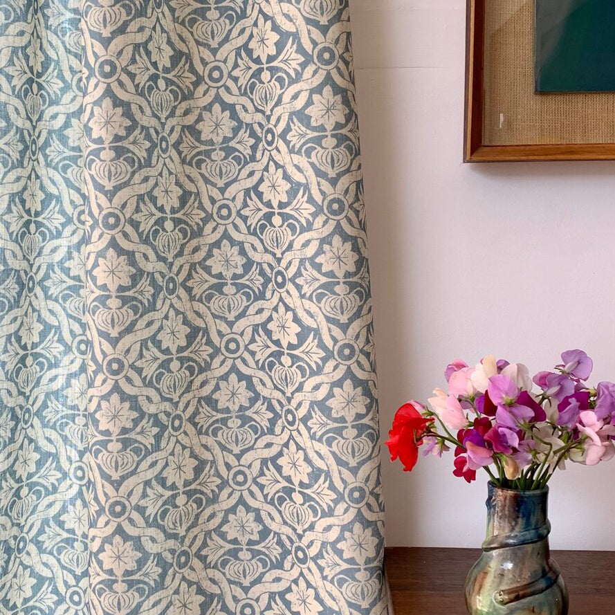 A curtain in a repeating blue and white floral grid pattern next to a wooden table with a vase of pink flowers on top.