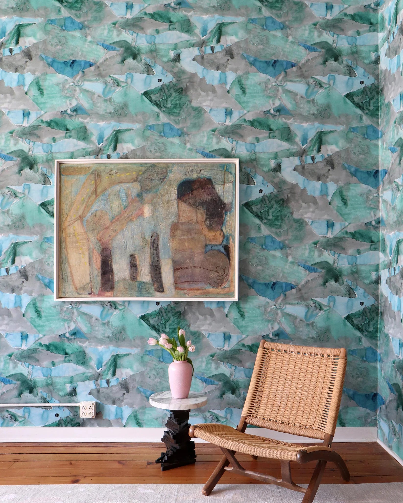 Room interior with a watercolor wallpaper in green and blue wall, wicker chair, and framed artwork.