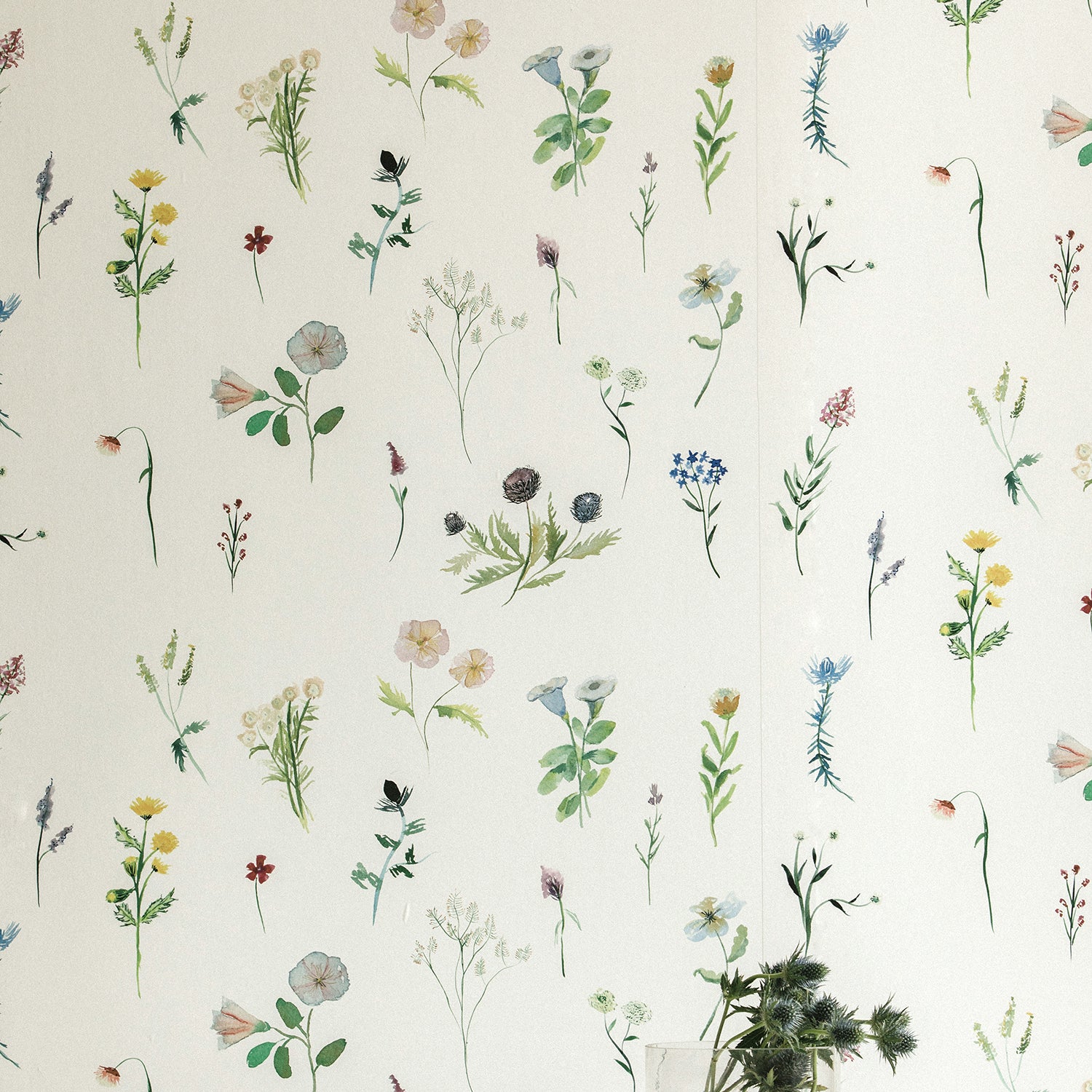 A vase of flowers stands in front of a wall covered in a painterly flower print in green and pastels on a cream field.