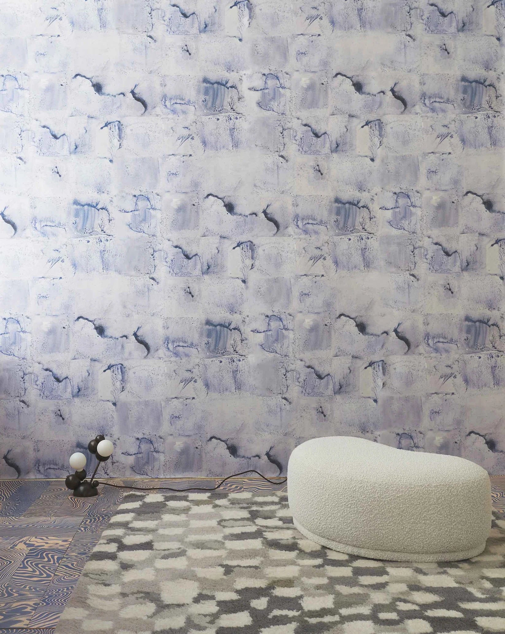 Room interior with a watercolor check wallpaper in blue featuring a white ottoman and small lamp.