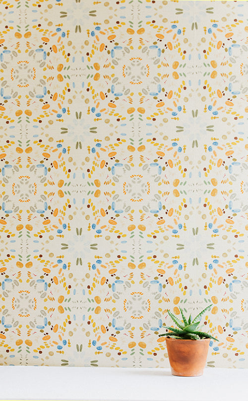 A potted plant stands in front of a wall covered in a painterly grid print in shades of blue and orange on a cream field.