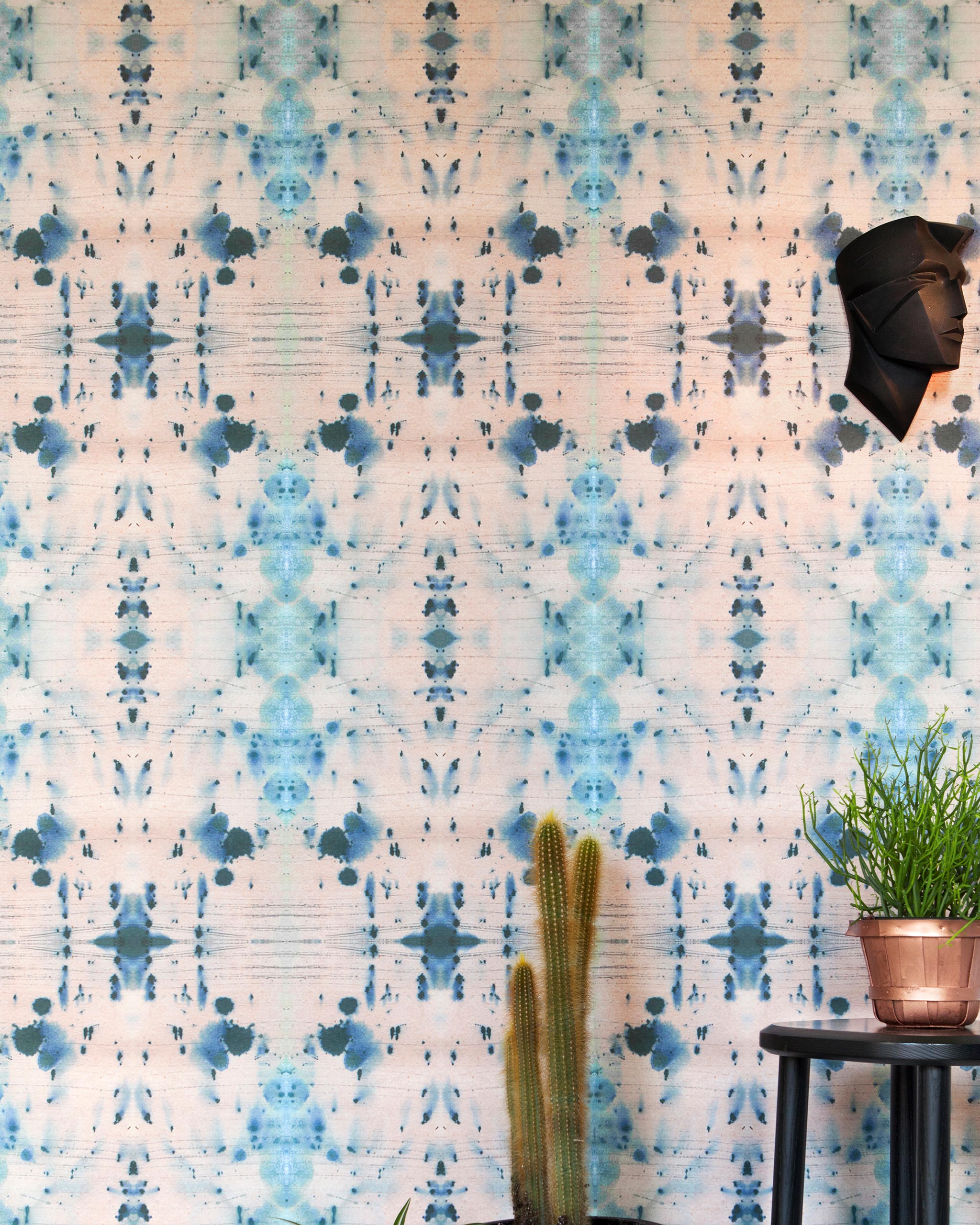 An end table and cactus stand in front of a wall papered in a watercolor ink blot print in blue, gray and light pink.