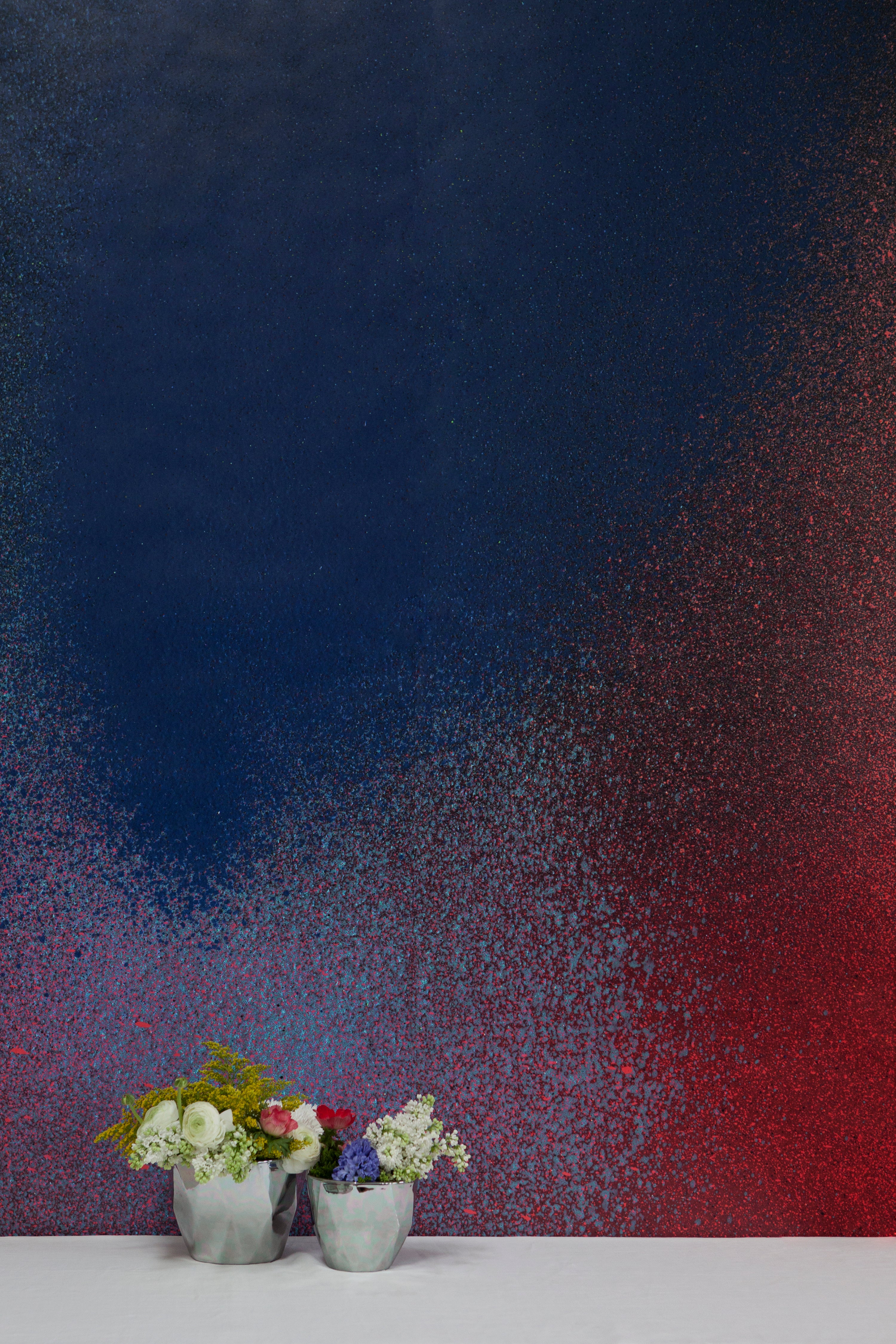 Two vases of flowers stand in front of a wall covered in an abstract ombré paint splatter print in red, blue and black.
