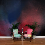 Chairs and a plant stand in front of a wall covered in an abstract ombré paint splatter print in red, blue and black.