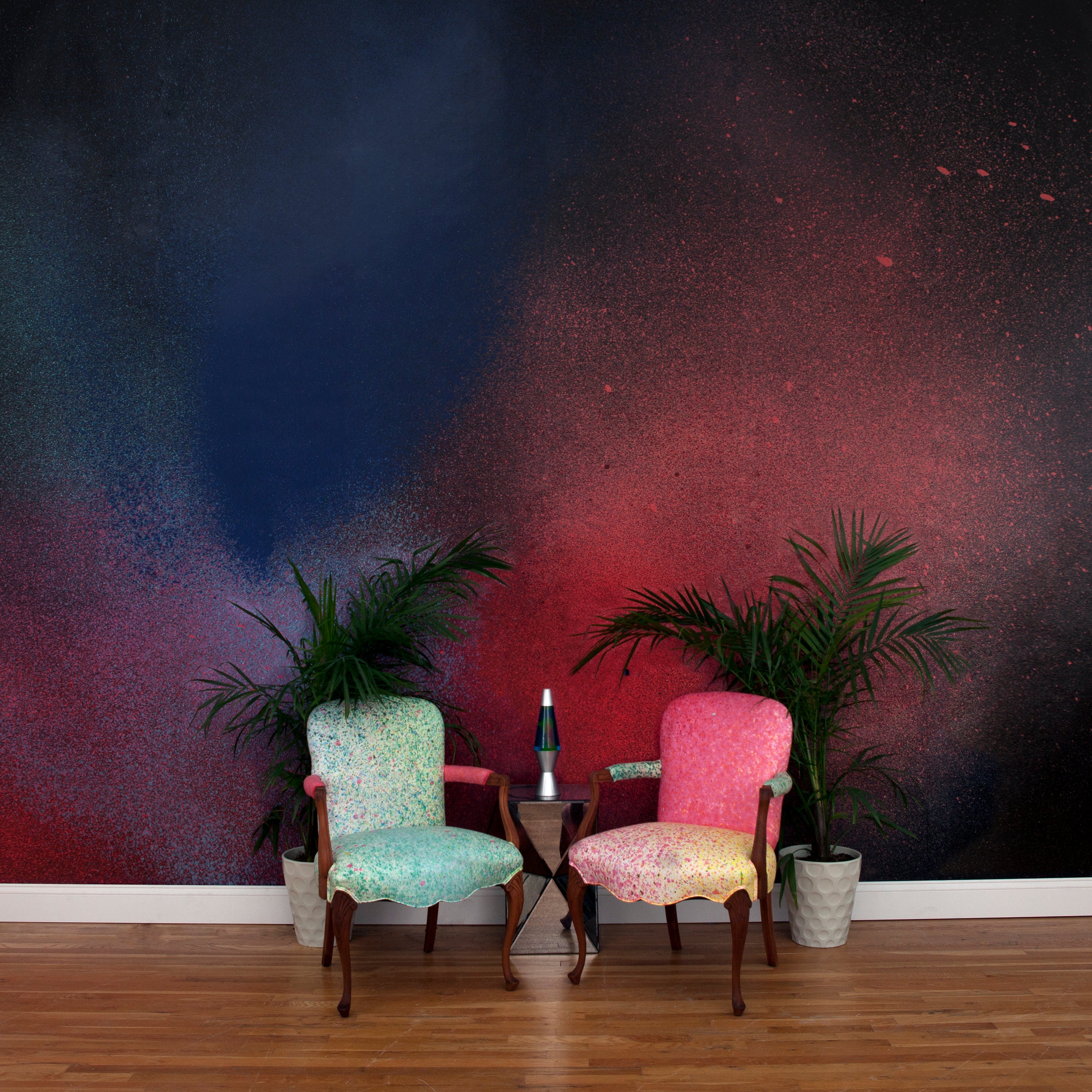 Chairs and a plant stand in front of a wall covered in an abstract ombré paint splatter print in red, blue and black.