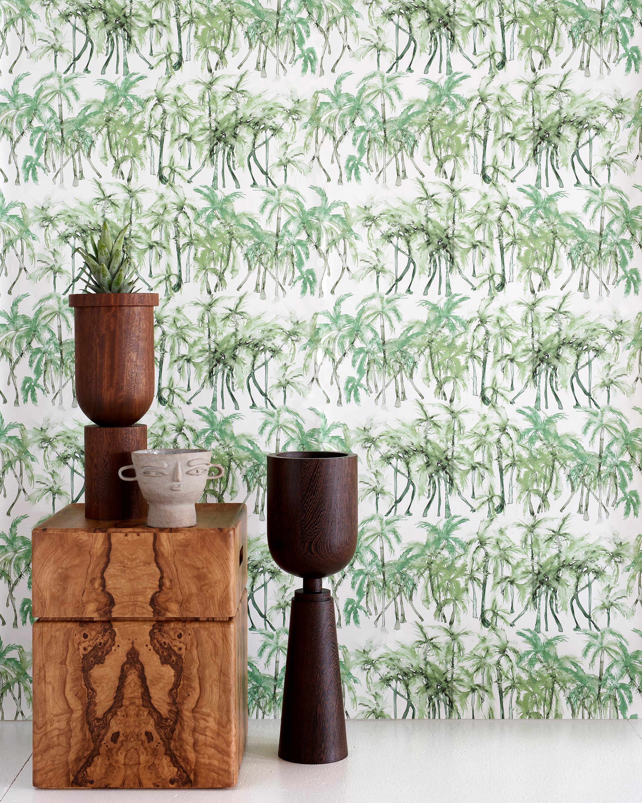 An end table with a plant and sculptures stands in front of a wall papered in a painterly palm tree print in green and white.