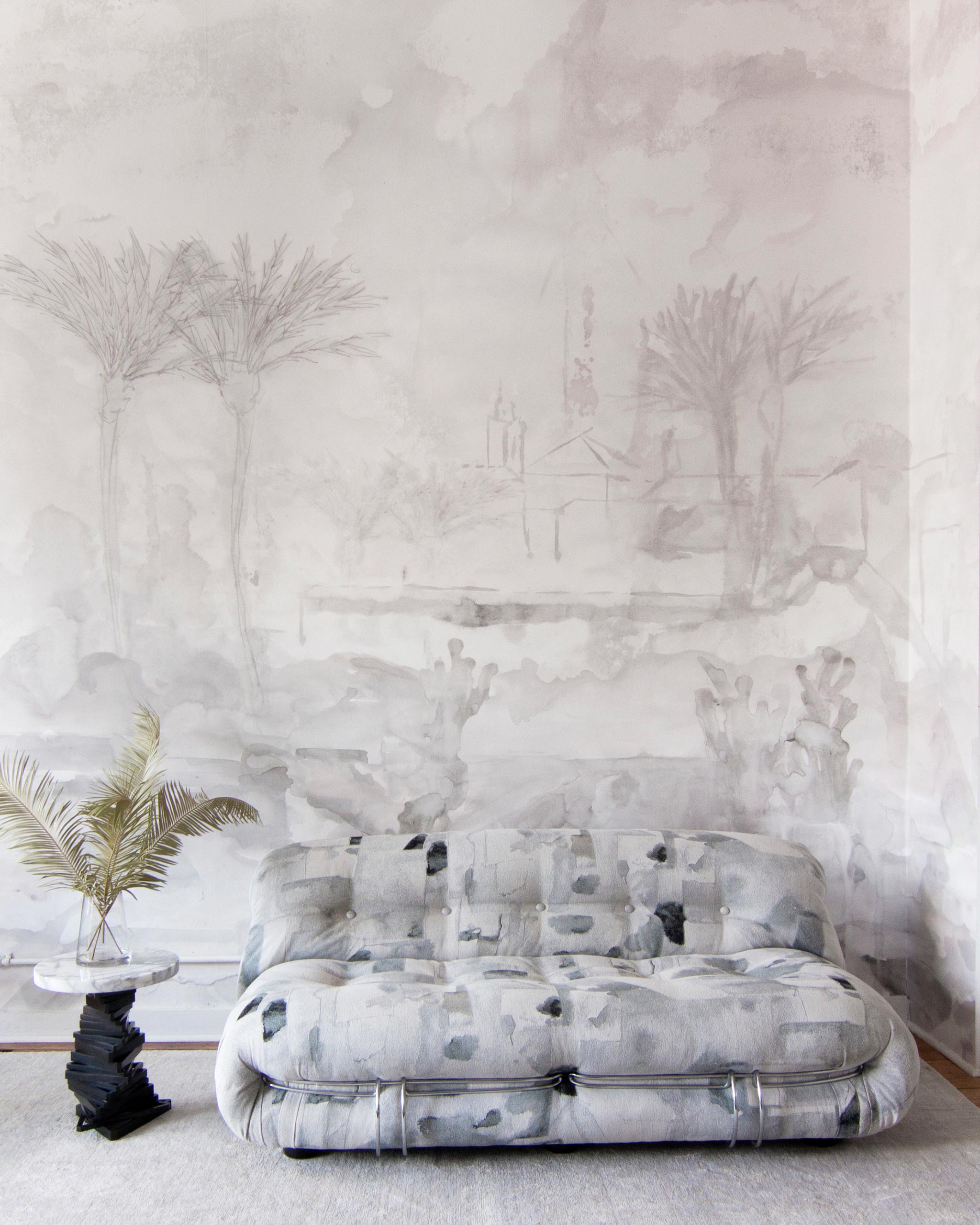 A modernist couch and end table stand in front of a wall papered in a watercolor painterly print in shades of gray and cream.