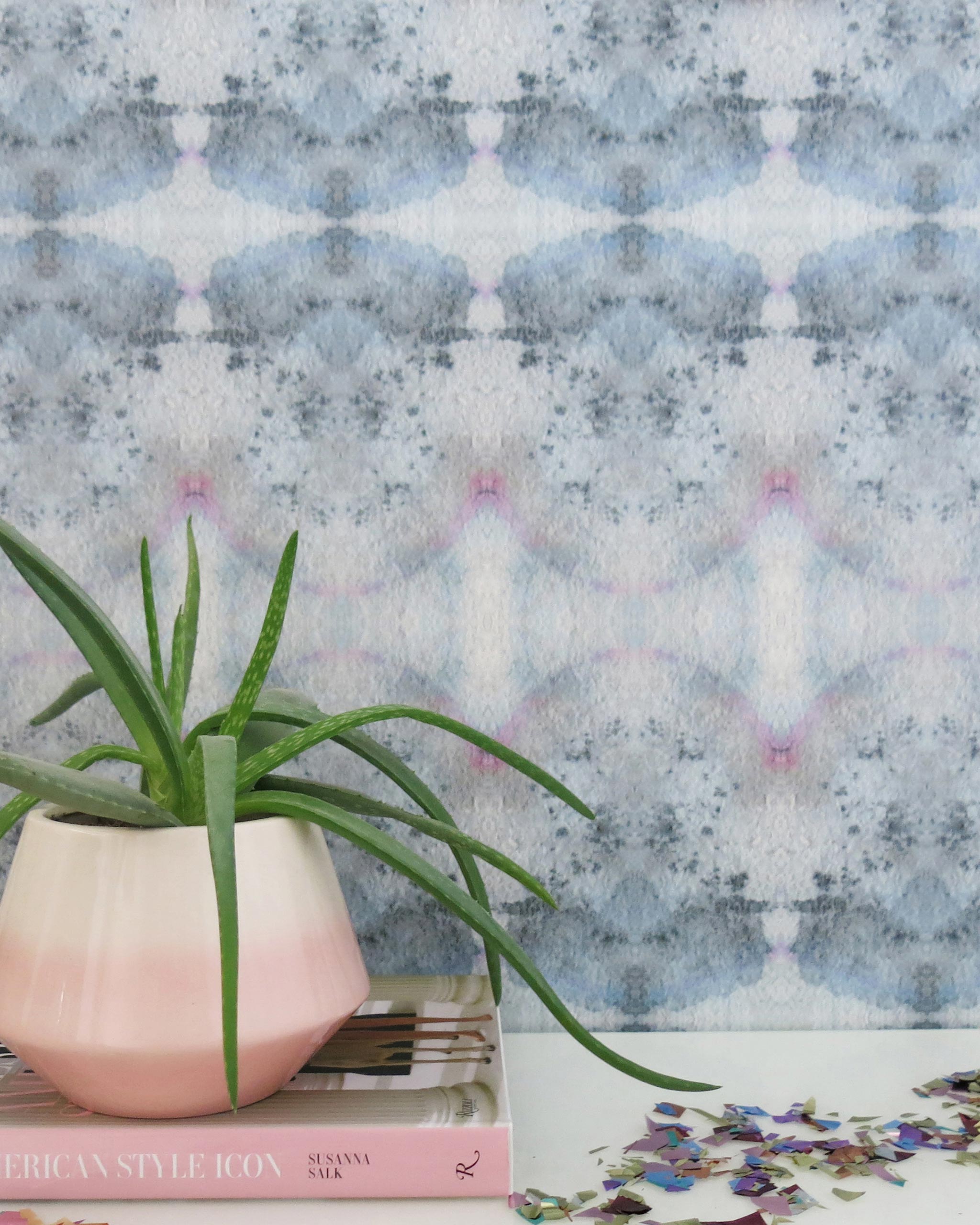A plant and book stand in front of a wall papered in a watercolor ink blot pattern in shades of gray, blue and pink.