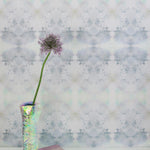 A vase with a flower stands in front of a wall papered in a watercolor ink blot pattern in shades of gray, green and pink.