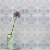 A vase with a flower stands in front of a wall papered in a watercolor ink blot pattern in shades of gray, green and pink.