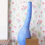 A modernist statue stands in front of a wall papered in a playful palm tree print in a rainbow of neon on a white field.