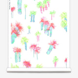 Partially unrolled wallpaper yardage in a playful palm tree print in a rainbow of neon shades on a white field.
